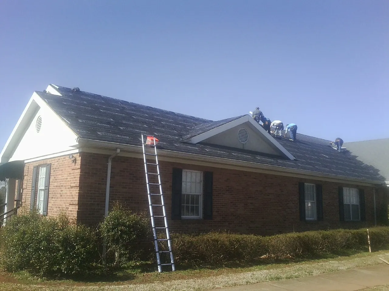 Crew tearing shingles off a brick ranch roof with a ladder set — Metal Roof Repair work by our crew in Amber
