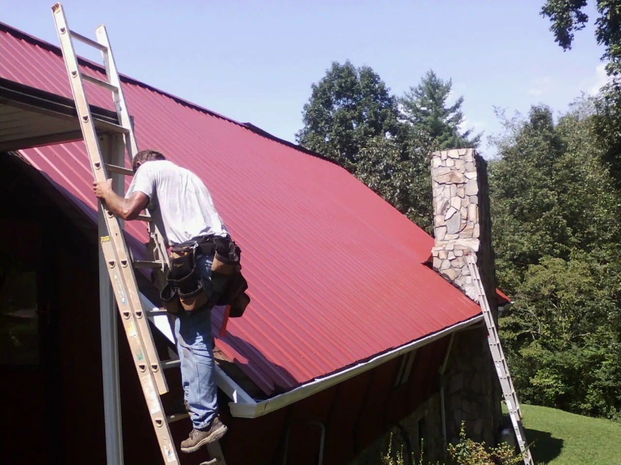 Roofer on a ladder beside a red metal roof and stone chimney — Soffit & Fascia Repair by Roofing Brothers in Amber